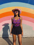 Bold patterned one-piece swimsuit with retro evil eye floral design, styled in festival-ready outdoor settings. Swimsuit features a scoop neckline, cheeky fit, and low scoop back. Ideal for summer vacations, pool parties, raves, and alt girl fashion. Made from stretchy, moisture-wicking fabric with UPF 50+ protection. Available in sizes XS to 3X. Colorful, statement swimwear designed for confident, maximalist style lovers.